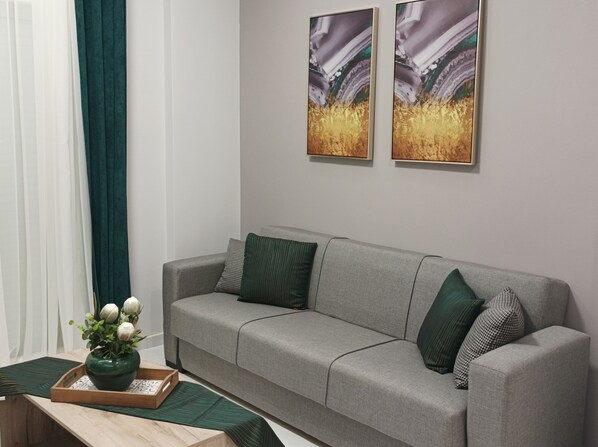 A modern living room with a grey sofa, green and patterned cushions, a wooden side table with a vase of flowers, and abstract paintings on the wall.