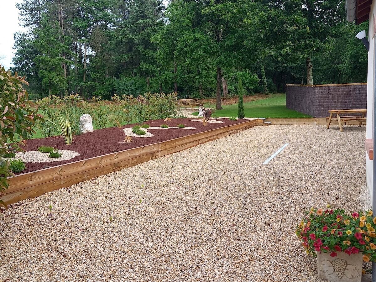Un camino de grava conduce a una terraza de madera con un banco, rodeada de jardines cuidados y árboles.