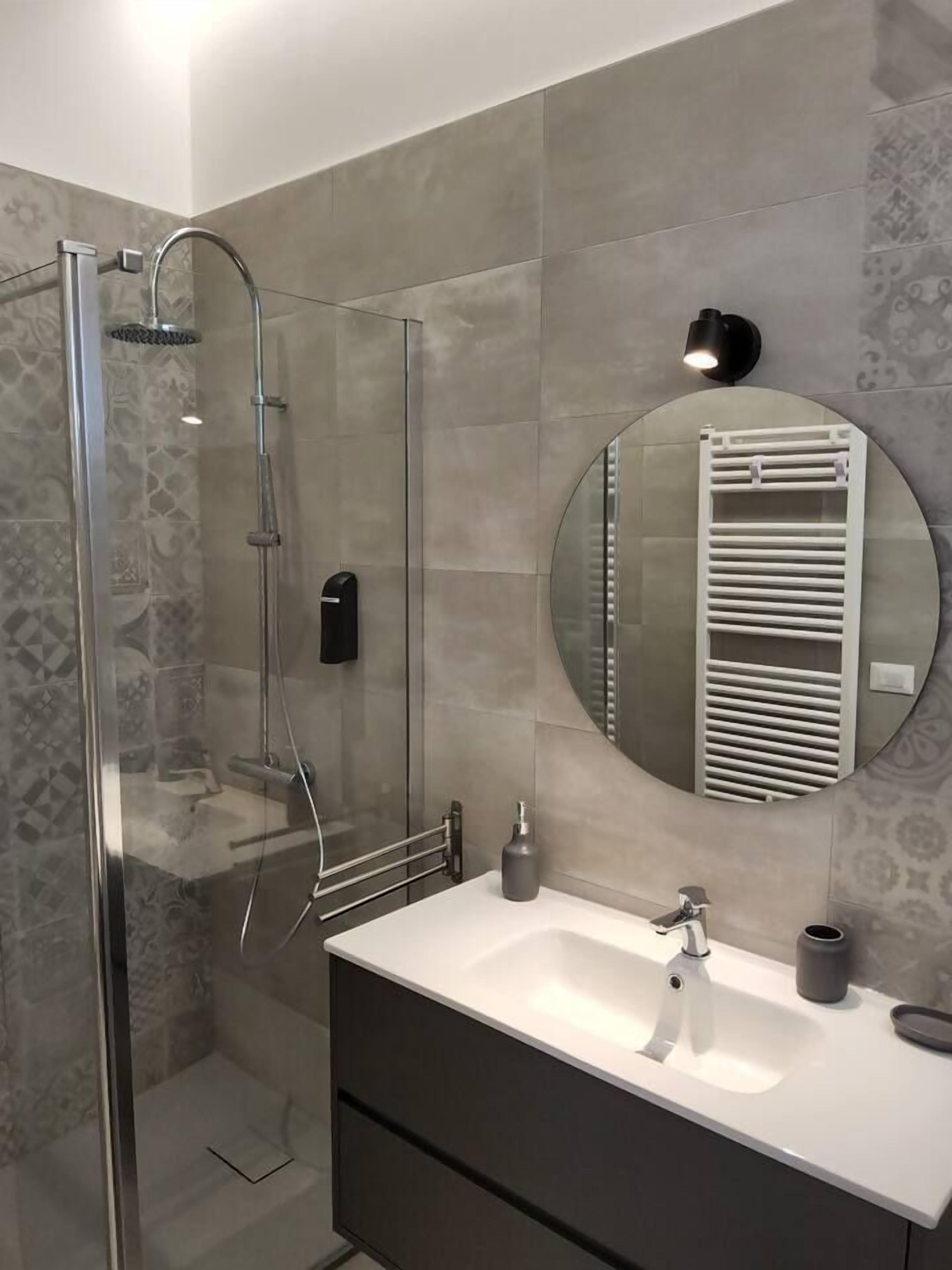 A modern bathroom with a glass-enclosed shower, a round mirror, and a white sink with a dark cabinet.