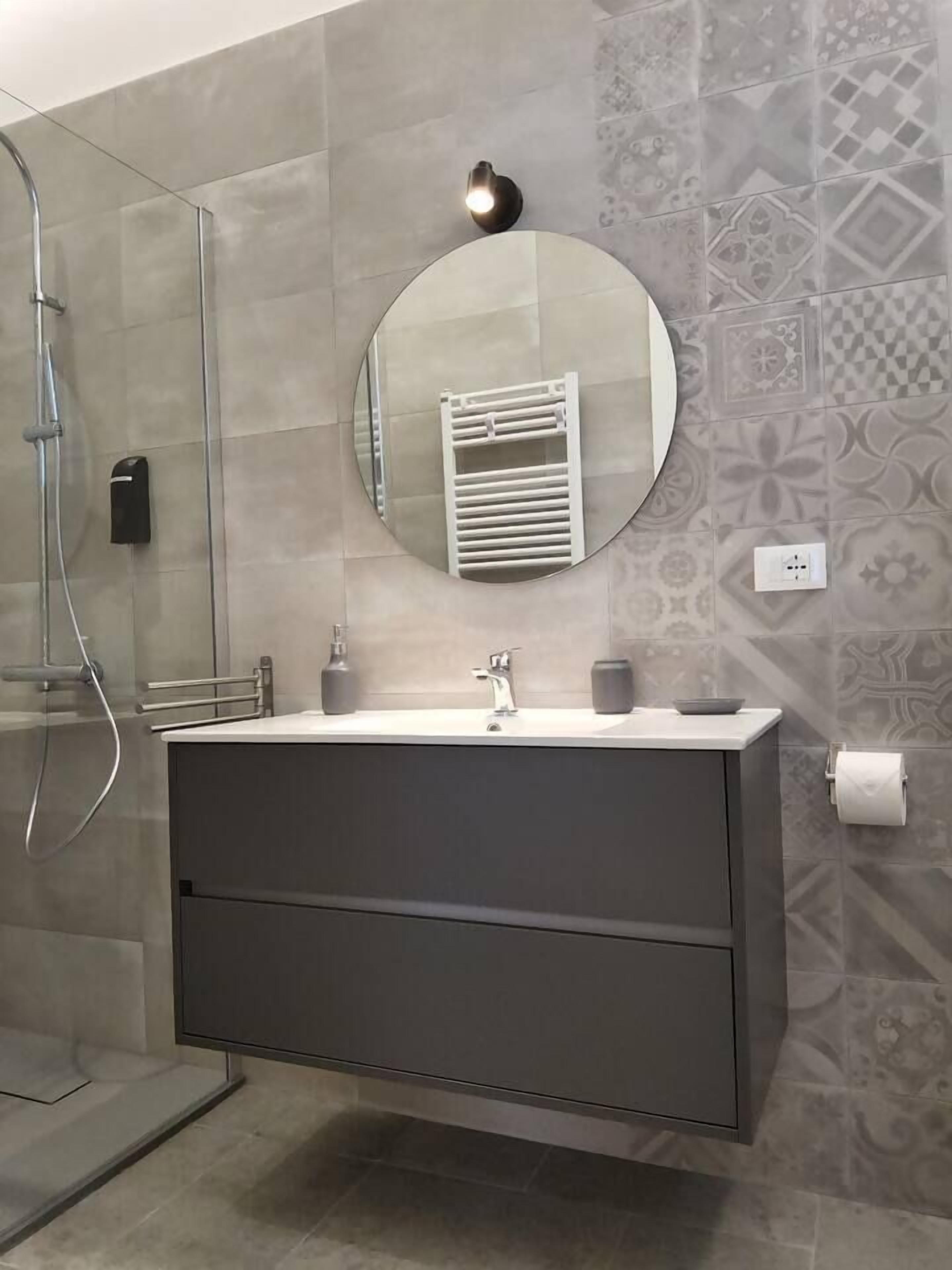 A modern bathroom with a glass shower enclosure, a round mirror, a white towel rack, and a grey vanity with a sink.