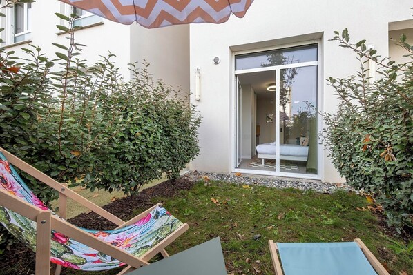 A patio with a wooden chair, a small table, and a large umbrella. There is a glass door leading to a room with a bed and a window. - Appart Hôtel Toulouse Zenith (Toulouse)