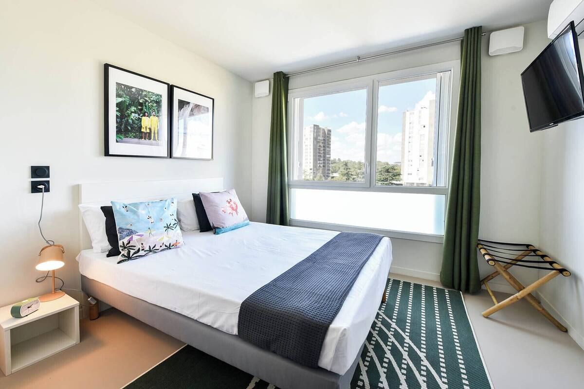 A hotel room with a bed, a side table with a lamp, a TV, a window with curtains, and a view of buildings.