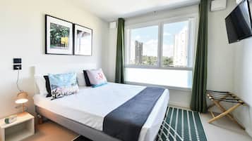 A hotel room with a bed, a side table with a lamp, a TV, a window with curtains, and a view of buildings.