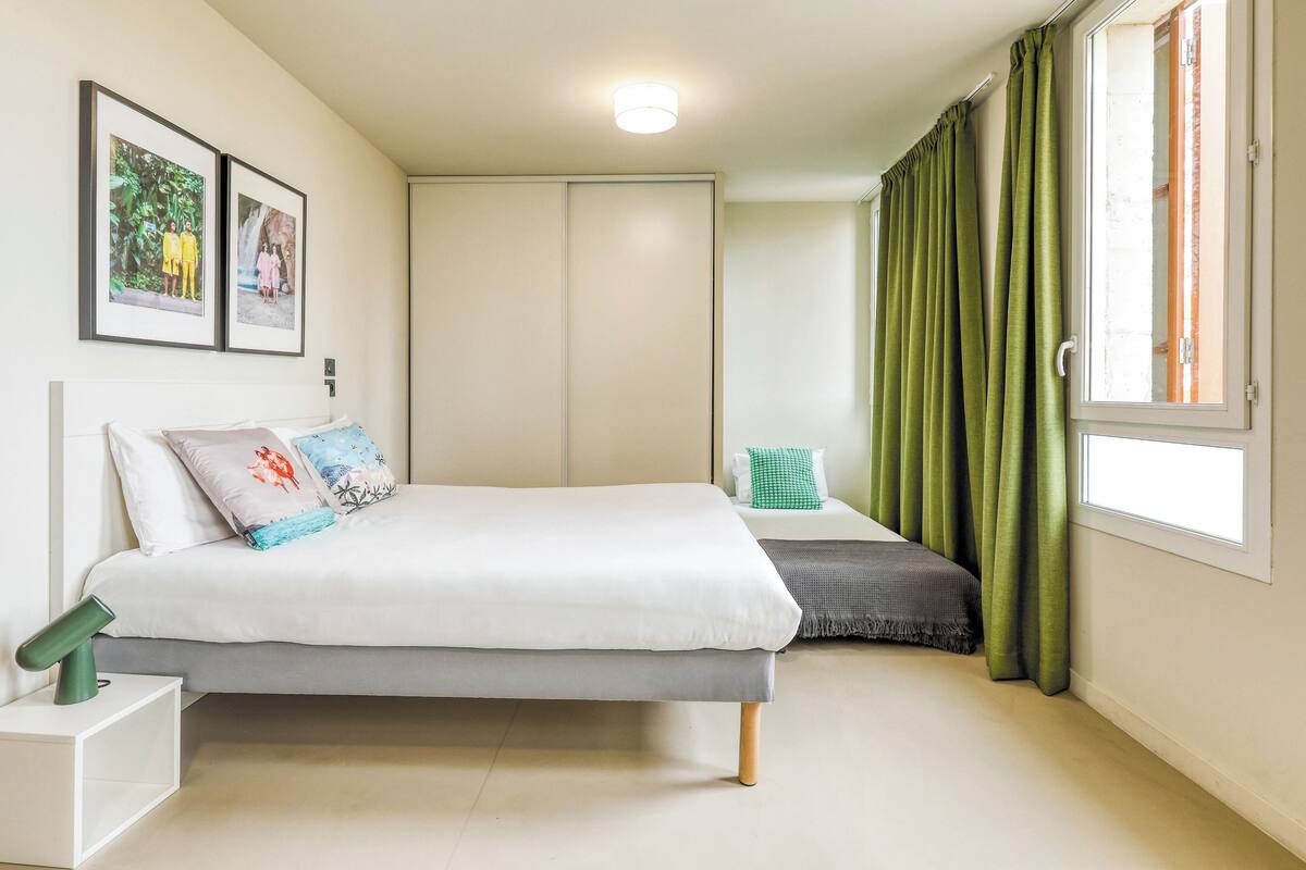 A bedroom with a bed, a nightstand, a wardrobe, a window with curtains, and two framed pictures on the wall.