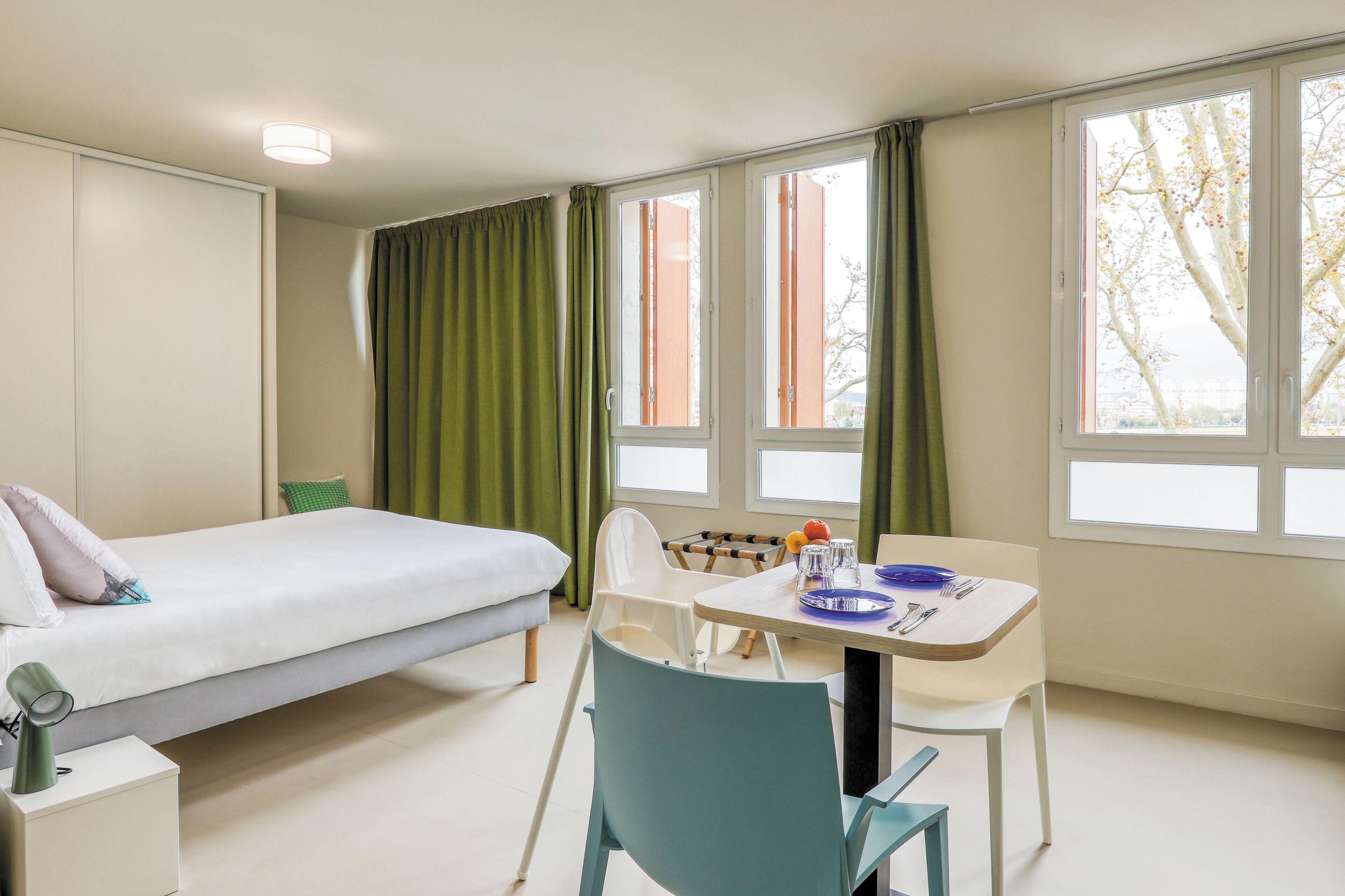 Una habitación con cama, una mesa de comedor con sillas y ventanas amplias con cortinas.