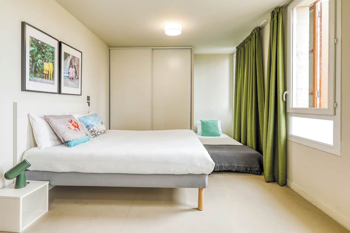 A bedroom with a bed, a nightstand, a wardrobe, a window with curtains, and two framed pictures on the wall.