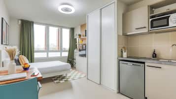 A compact room with a bed, a desk, a microwave, a dishwasher, and a window with curtains.