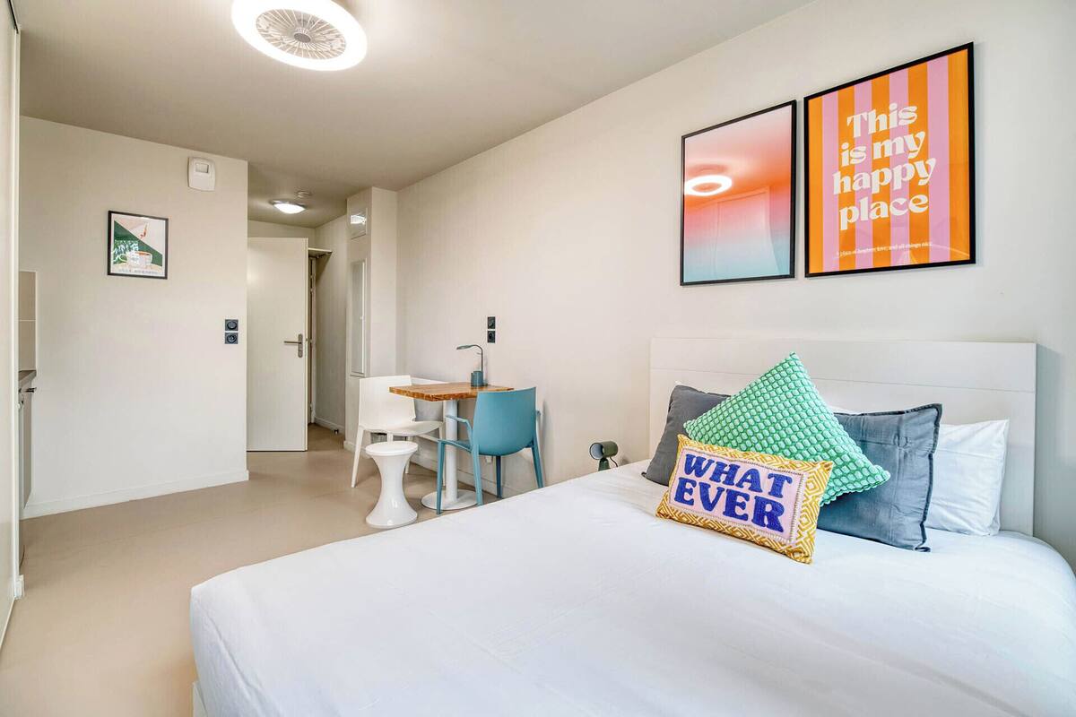 A hotel room with a bed, a desk, a chair, and two framed pictures on the wall.