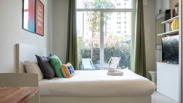 Un dormitorio con cama, almohadas, una ventana con cortinas, vistas a edificios y una pequeña mesa con una silla.