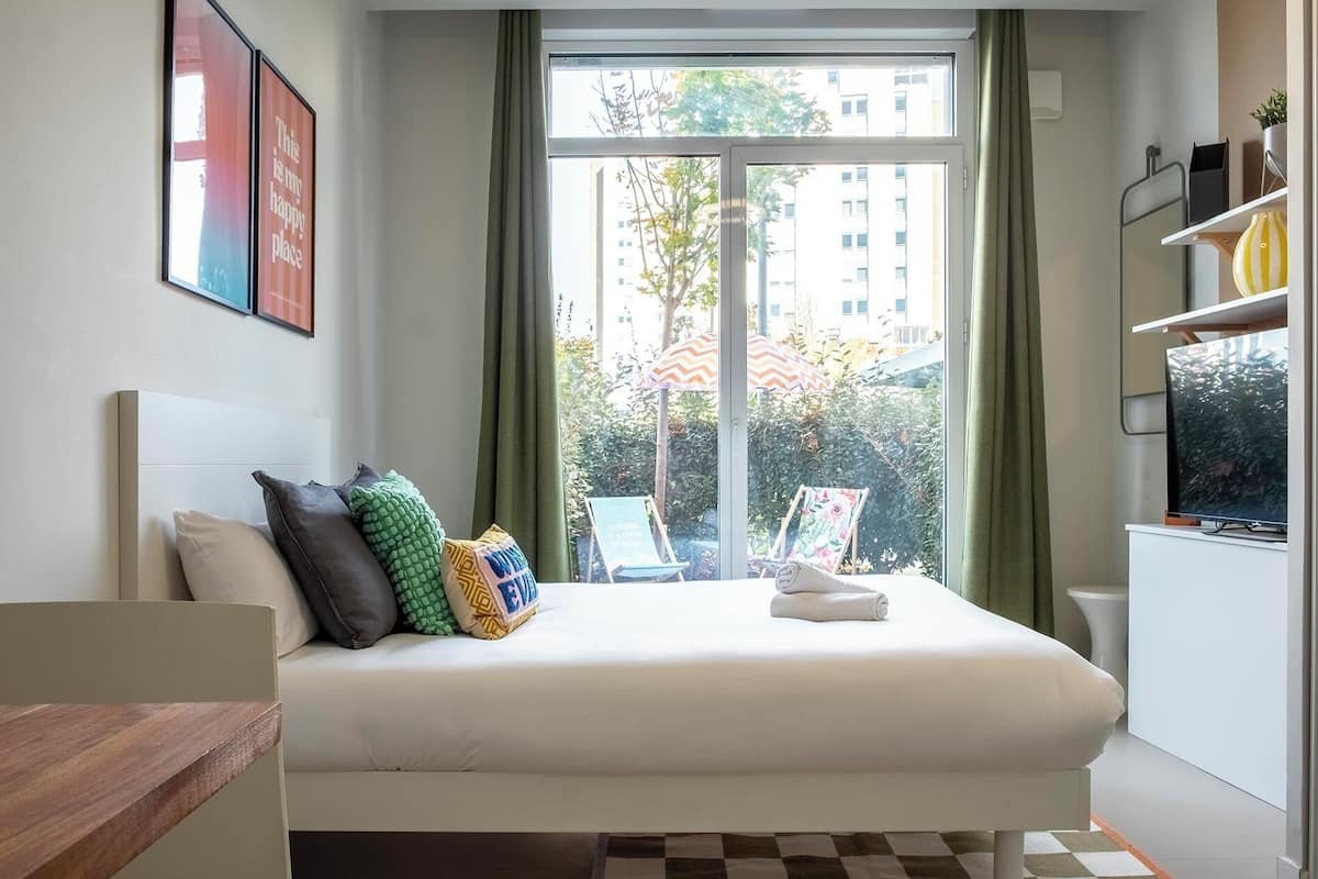 A bedroom with a bed, pillows, a window with curtains, a view of buildings, and a small table with a chair.