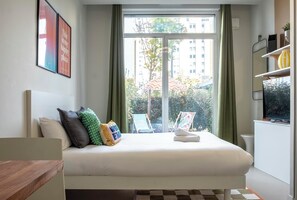 A bedroom with a bed, pillows, a window with curtains, a view of buildings, and a small table with a chair. - Appart Hôtel Toulouse Zenith (Toulouse)