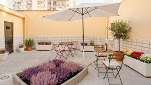 Une terrasse extérieure avec une table, des chaises, un parasol, des plantes en pot et un parterre de fleurs.