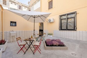 Une terrasse avec une table et des chaises, un parasol blanc et un jardinier orné de fleurs violettes.