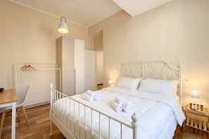 A bedroom with a white metal bed frame, a wooden desk, a chair, and a hanging rack with clothes.