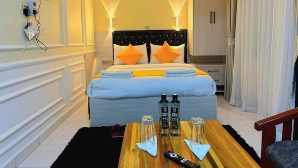 A hotel room with a bed, a TV mounted on the wall, a wooden table with bottles and a remote control, and a wardrobe. - Bijou Hotel Mlolongo (Mlolongo)