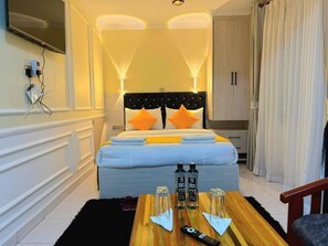 Quarto de hotel com cama, televisão fixada na parede, mesa de madeira com garrafas e um comando remoto, e um guarda-roupa.