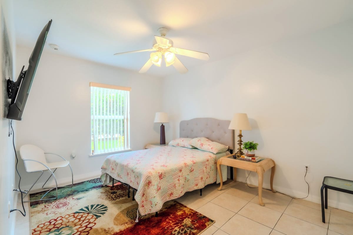 A bedroom with a bed, two bedside tables, a ceiling fan, a window, a chair, and a TV mounted on the wall.