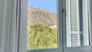 Ett fönster med vita persienner, utsikt över ett bergigt landskap och ett grönt träd i förgrunden.