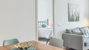 Ett modernt vardagsrum med ett matbord i trä, en grå soffa och en säng med ett vitt täcke och en turkosfärgad kudde.