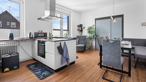 A modern kitchen with a central island, a dining area with a grey sofa and chairs, and a large window providing natural light.