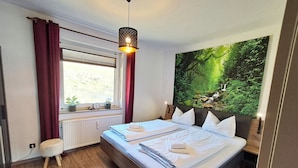 A bedroom with two beds, a window with curtains, a radiator, a small stool, and a large forest-themed wall hanging.