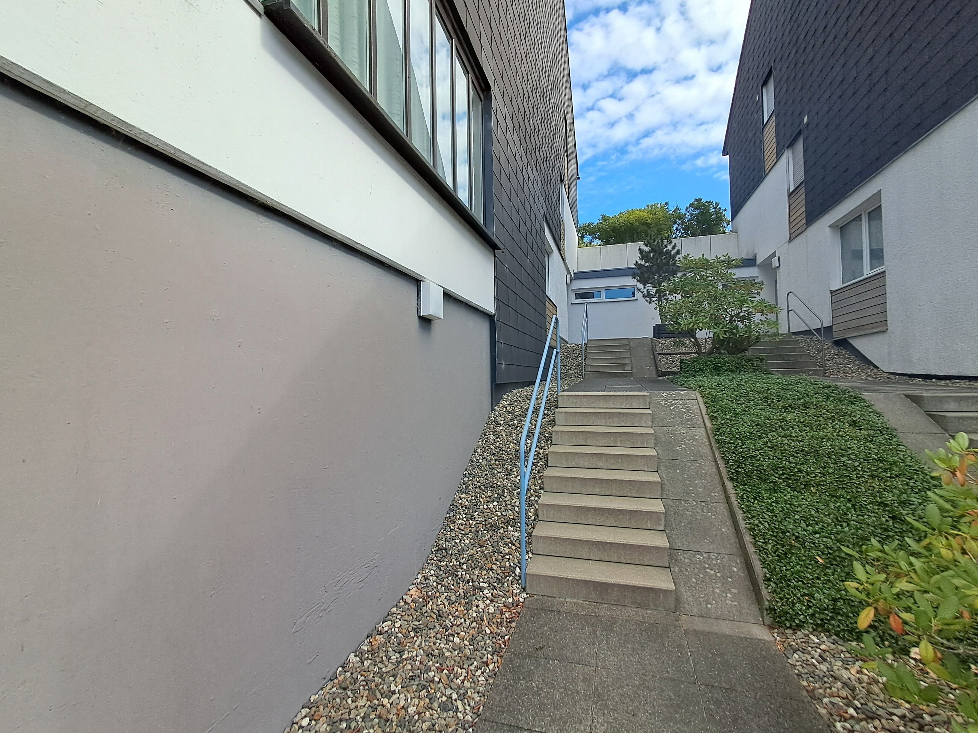 Un sendero estrecho que conduce a un edificio de diseño moderno, con una escalera de concreto y una barandilla de metal.