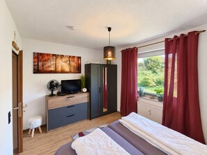 Une chambre à coucher comprenant un lit, une commode, un téléviseur, un ventilateur, une horloge et une fenêtre avec des rideaux.