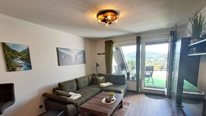 A modern living room with a sofa, coffee table, and a TV. There are two paintings on the wall and a balcony with a view of the outdoors.