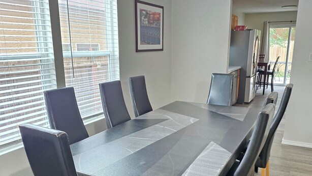 Een vergaderruimte met een grote tafel, zwarte stoelen en houten poten. Er zijn ramen met zonwering, een ingelijste foto aan de muur en een kitchenette op de achtergrond.