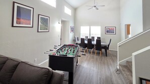 Een ruime woonkamer met een tafelvoetbaltafel, een eettafel met stoelen en ingelijste foto’s aan de wand.
