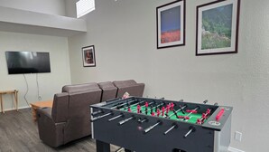 Een kamer met een tafelvoetbalspel, een aan de muur bevestigde televisie, een bank en ingelijste foto's.