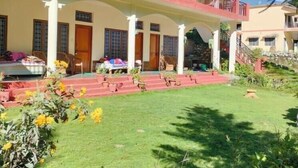 A two-story building with a covered patio area, multiple balconies, and a well-maintained garden with various plants and flowers. - Anand-Ganga Yoga Guesthouse (Bhatwari)