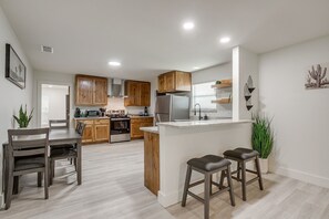 A modern kitchen with wooden cabinets, a central island, bar stools, and a dining table. - 4BR · Sleeps 9 · Walk to Tarleton · Fenced Yard (Stephenville)