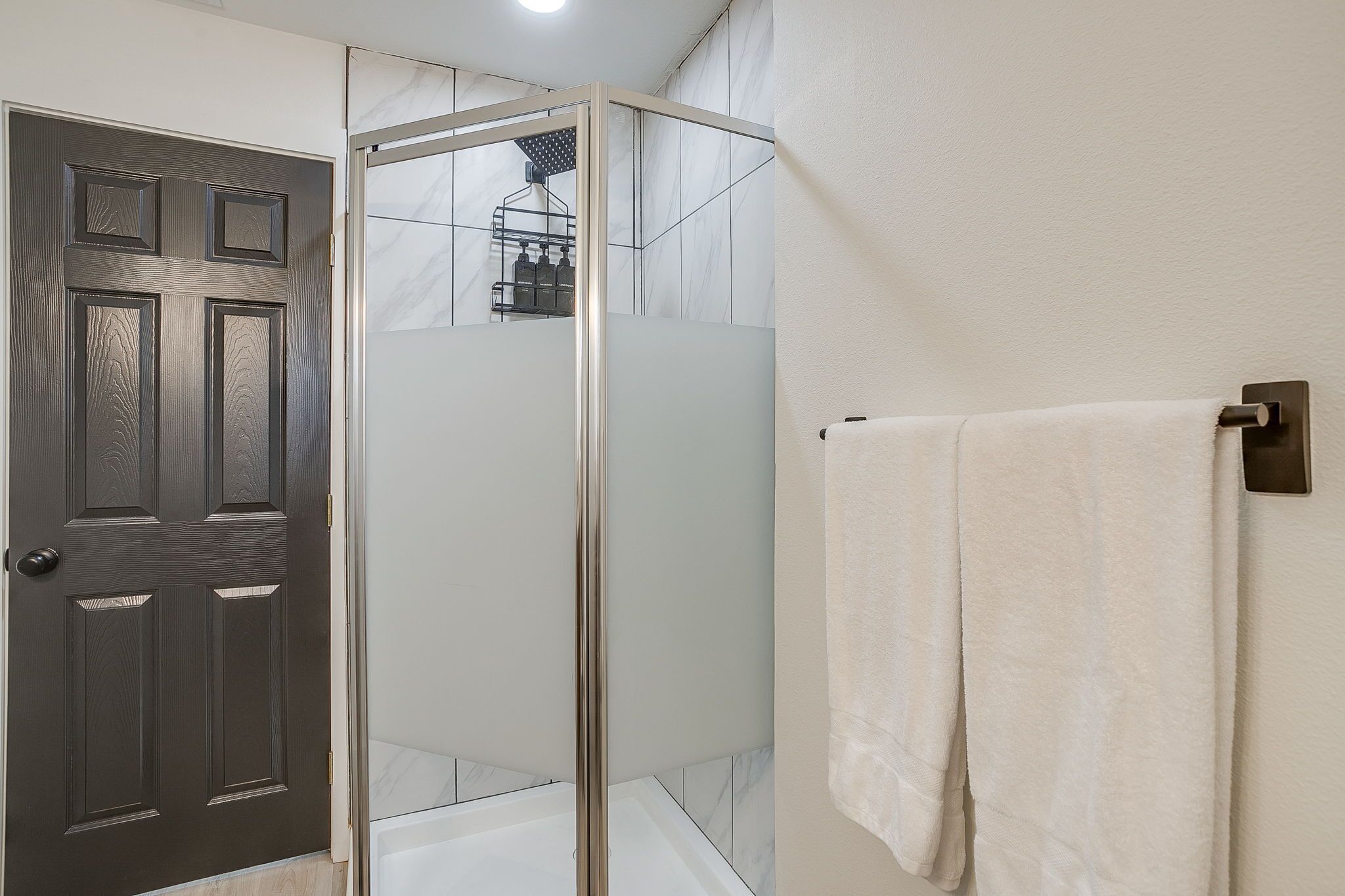 A bathroom with a glass-enclosed shower, a white towel hanging on a wall-mounted towel rack, and a dark wooden door.