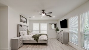 A bedroom with a bed, nightstands, a dresser, a ceiling fan, a TV, and large windows with blinds.