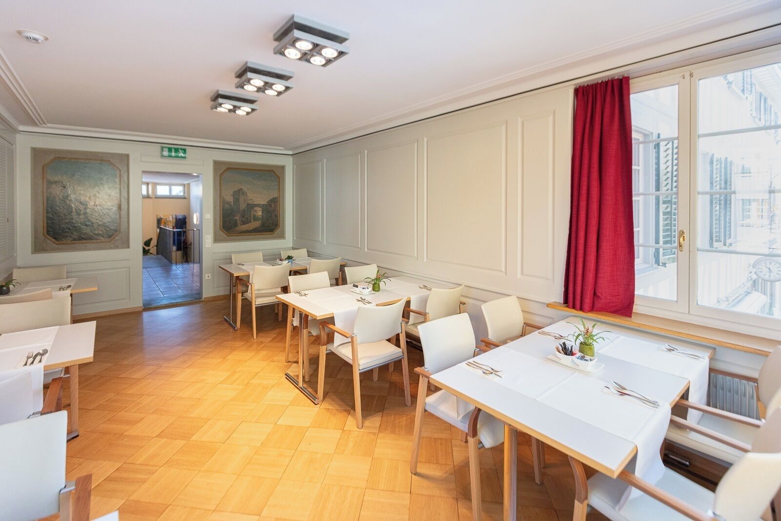 Un comedor con mesas y sillas blancas, una cortina roja y vista a la ciudad a través de la ventana.
