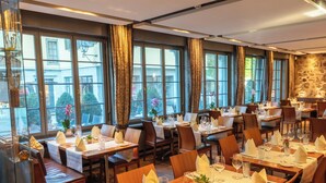 Un restaurante con varias mesas dispuestas para comer, ventanas grandes con cortinas y una pared de piedra al fondo.