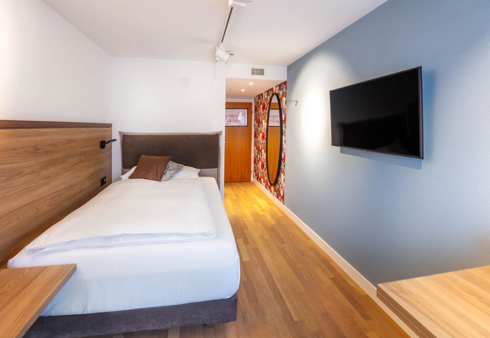 A modern hotel room with a wooden headboard, a large bed with white linens, a flat-screen TV mounted on the wall, and a wooden floor.