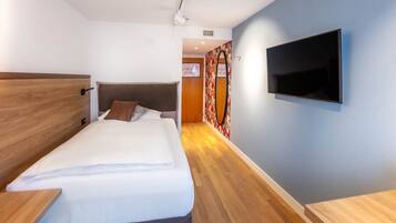 A modern hotel room with a wooden headboard, a large bed with white linens, a flat-screen TV mounted on the wall, and a wooden floor.