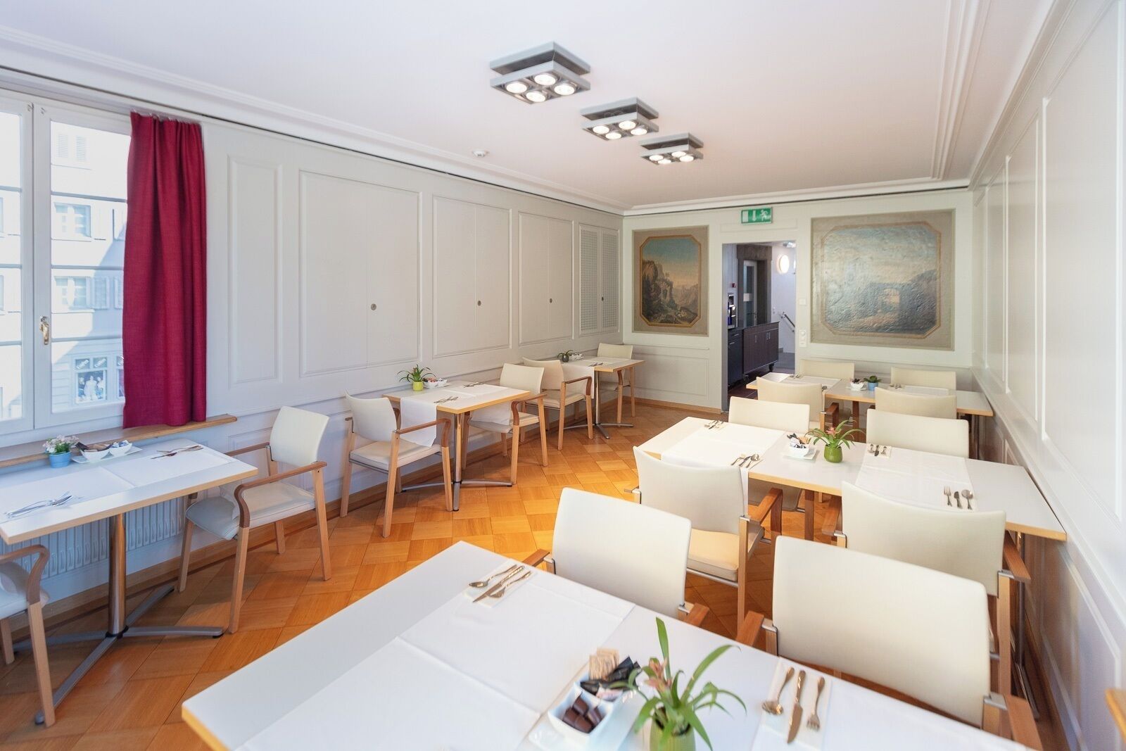 Un espace repas comprenant plusieurs tables et chaises, une fenêtre ornée d’un rideau rouge et des œuvres d’art accrochées aux murs.