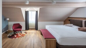 Un dormitorio moderno con suelo de madera, una cama grande con cabecera de madera, una silla roja, un banco y una ventana con cortinas.