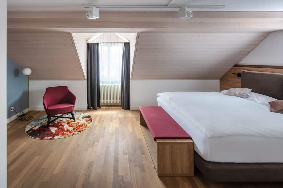A modern bedroom with a wooden floor, a large bed with a wooden headboard, a red chair, a bench, and a window with curtains.