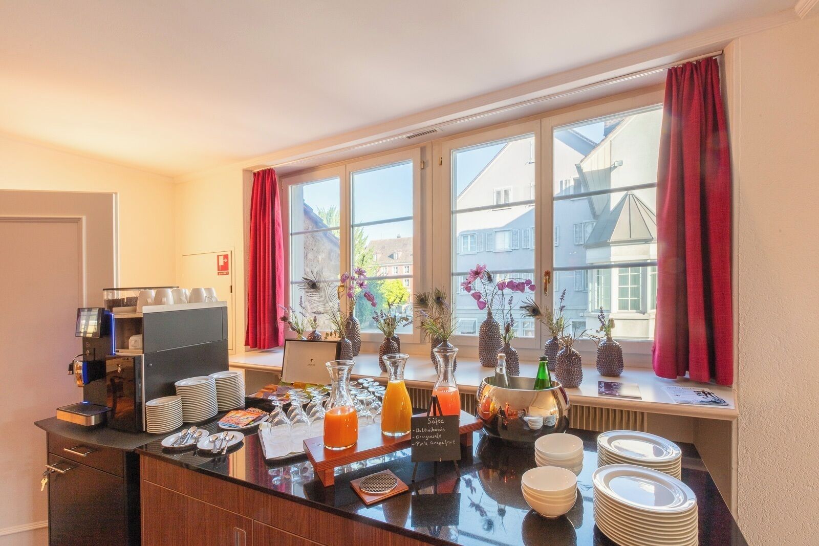 Une chambre d’hôtel équipée d’une kitchenette, d’une machine à café, de produits pour le petit-déjeuner et offrant une vue sur les bâtiments à travers de grandes fenêtres.