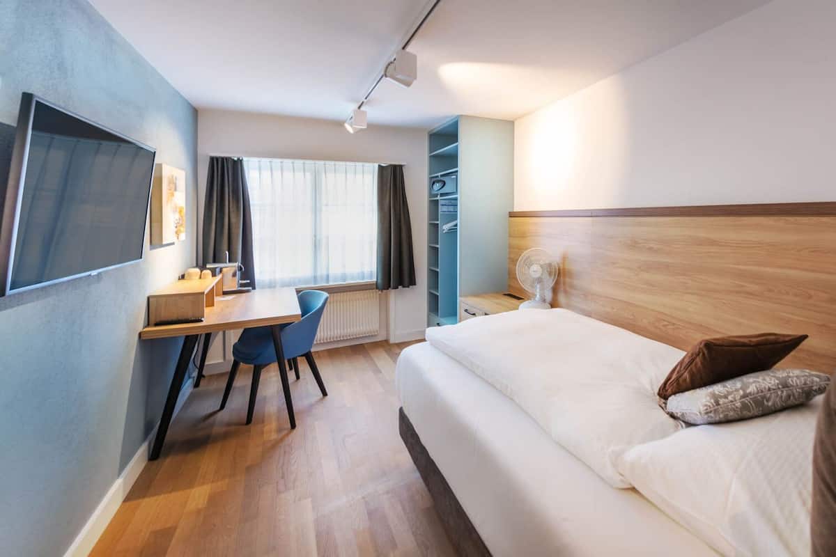 A hotel room with a bed, a desk with a chair, a TV, and a window with curtains.