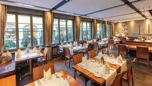 Un restaurante con varias mesas dispuestas para comer, ventanas grandes con cortinas y una pared de piedra con una lámpara moderna.