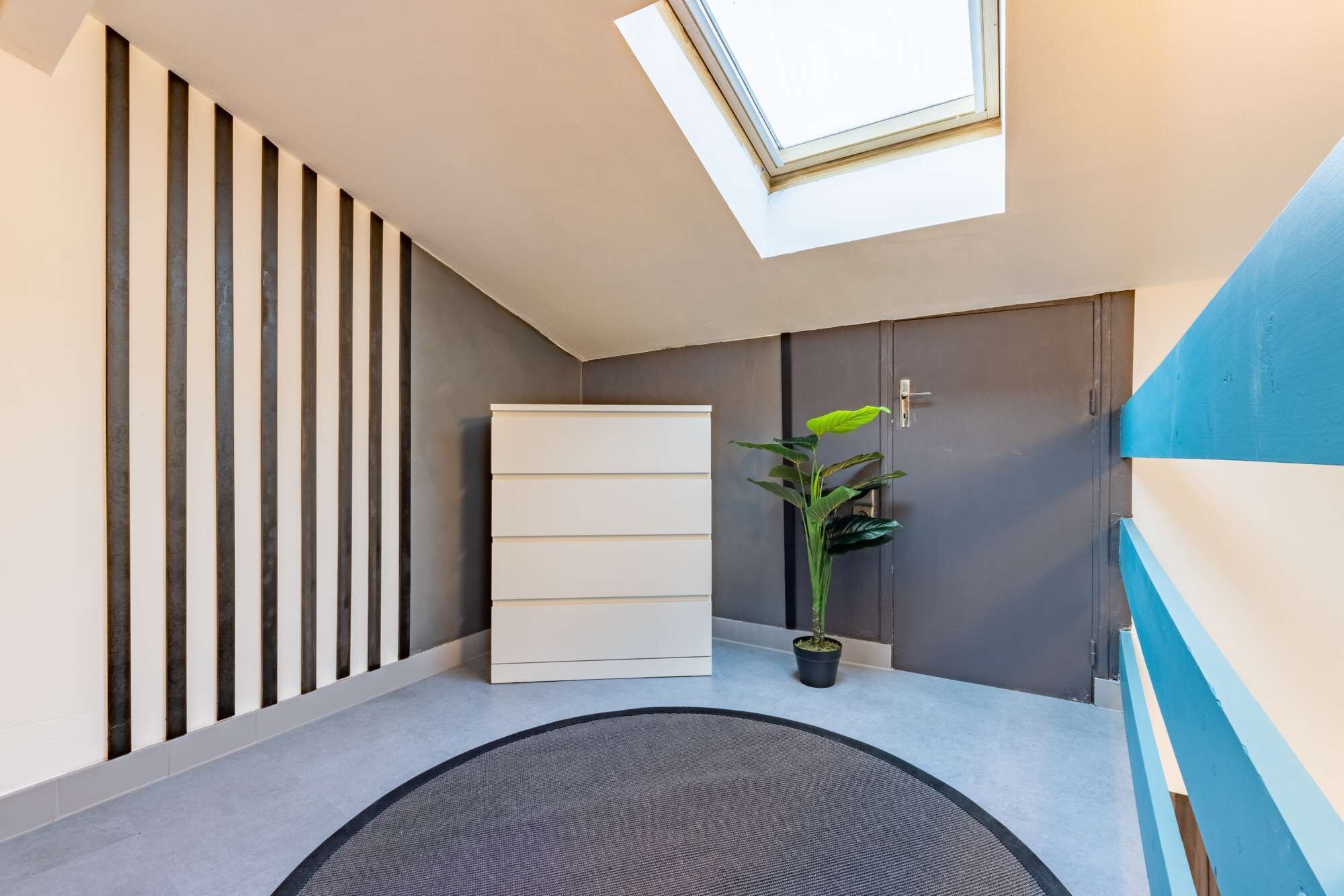 Un couloir moderne avec une lucarne, une plante en pot, un meuble blanc et un mur d’accent bleu.
