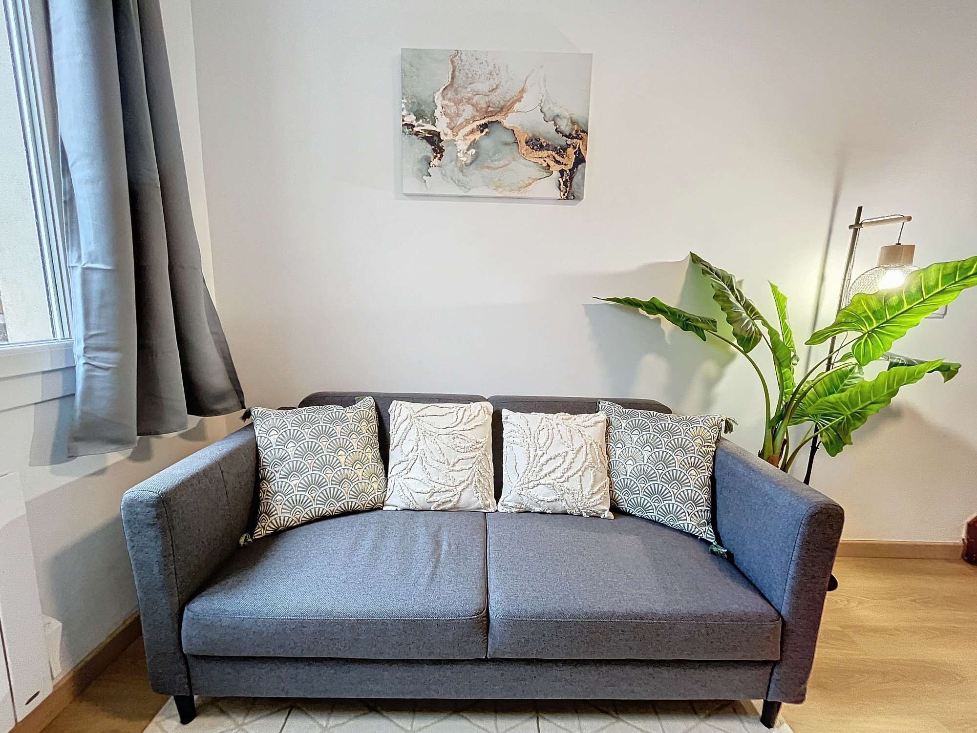 A grey sofa with patterned cushions, a wall-mounted abstract painting, and a large green plant.