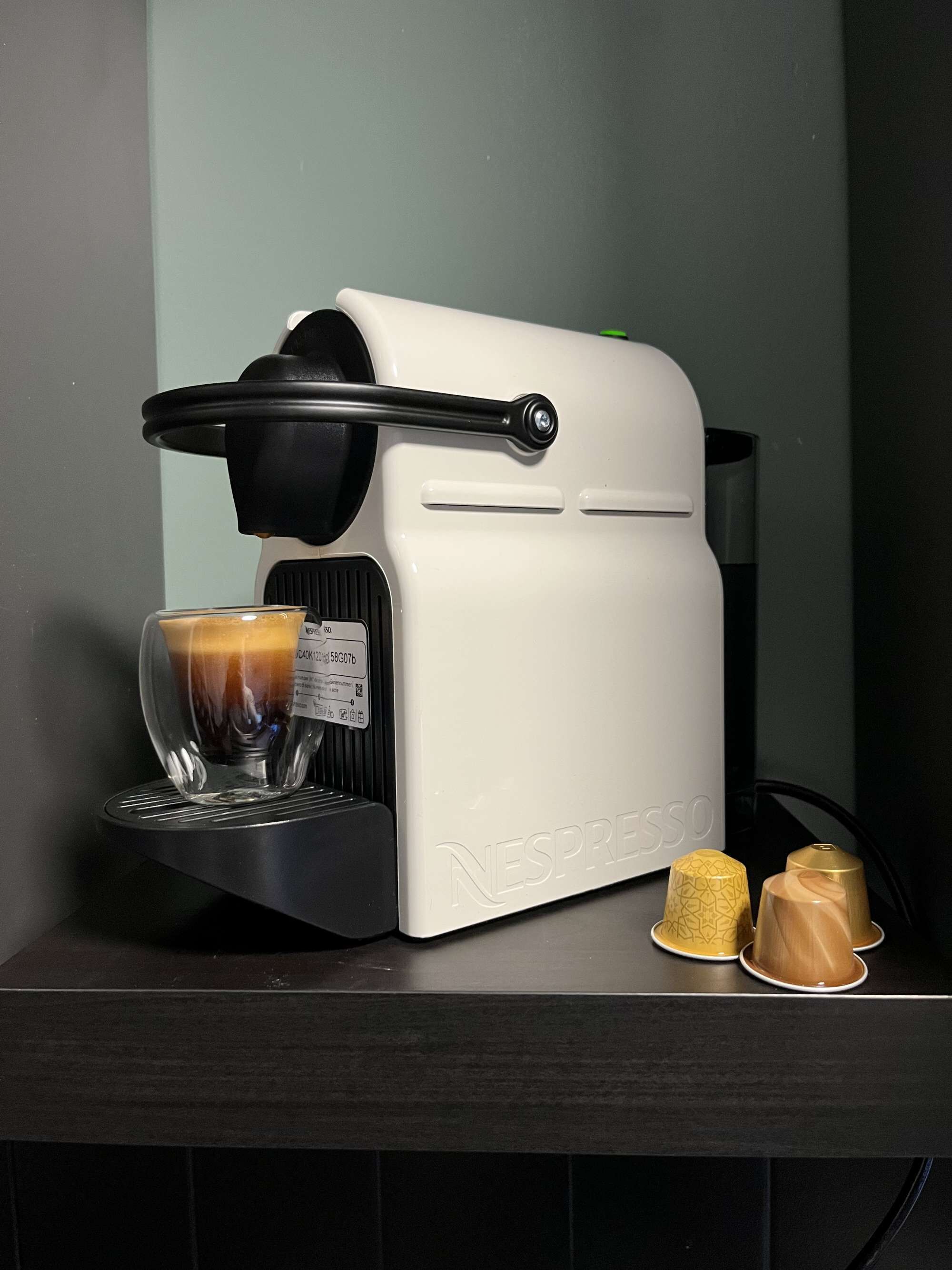 A Nespresso coffee machine with a glass cup of coffee, two capsules, and a black coffee grinder on a dark countertop.