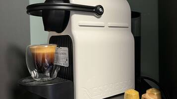 A Nespresso coffee machine with a glass cup of coffee, two capsules, and a black coffee grinder on a dark countertop.