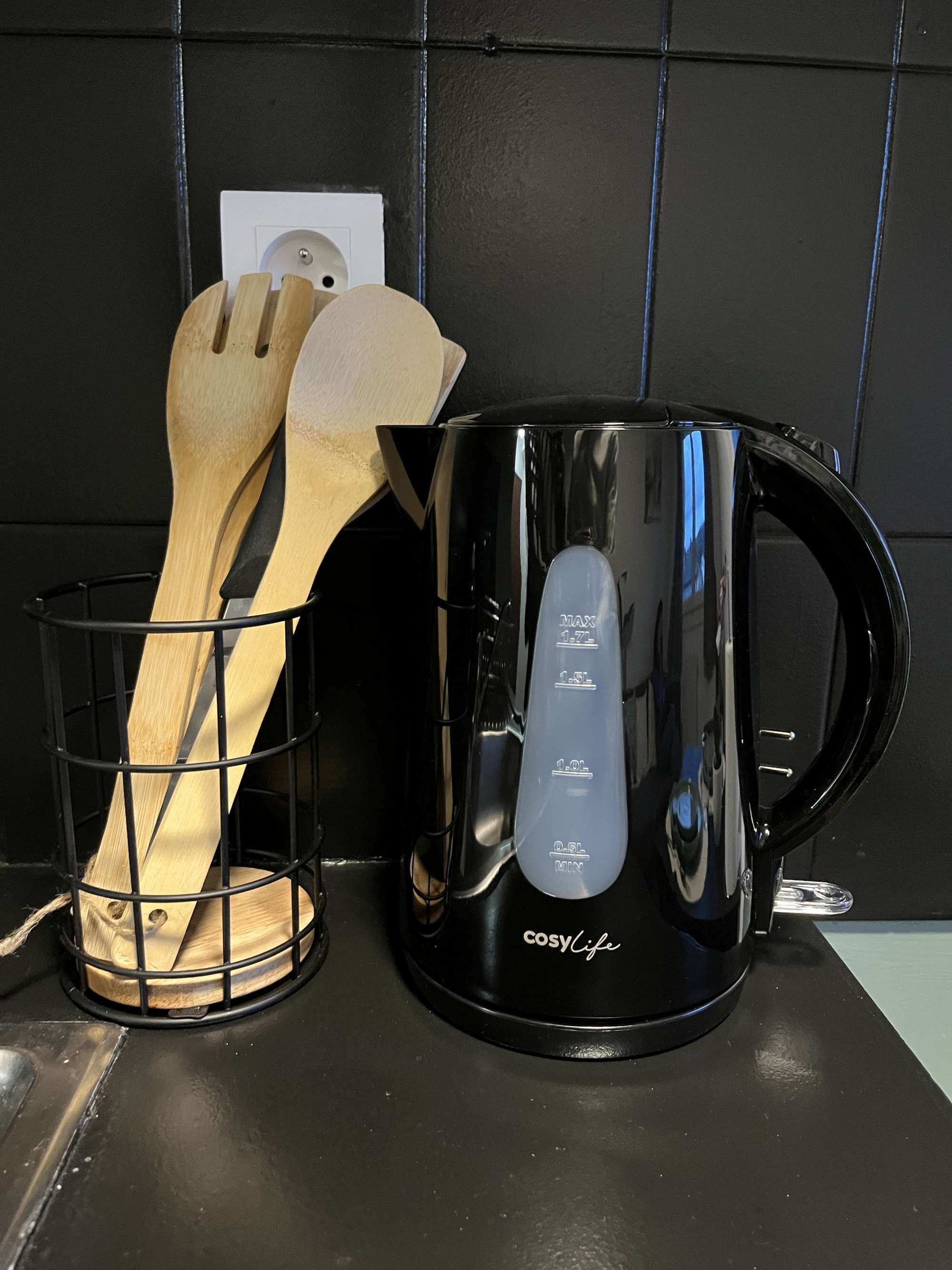 Een zwarte koffiezetapparaat met maatstrepen en houten lepels in een mand van metaaldraad op een keukentafel.